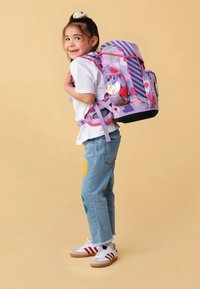 Un sac à dos violet avec des motifs colorés, des poches latérales en maille et un badge circulaire. Porté par un enfant portant une chemise blanche et un jean bleu.
