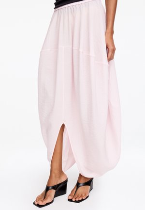Femme portant une jupe taille haute rose clair avec une fente à l'avant et des sandales compensées noires à bouts carrés.