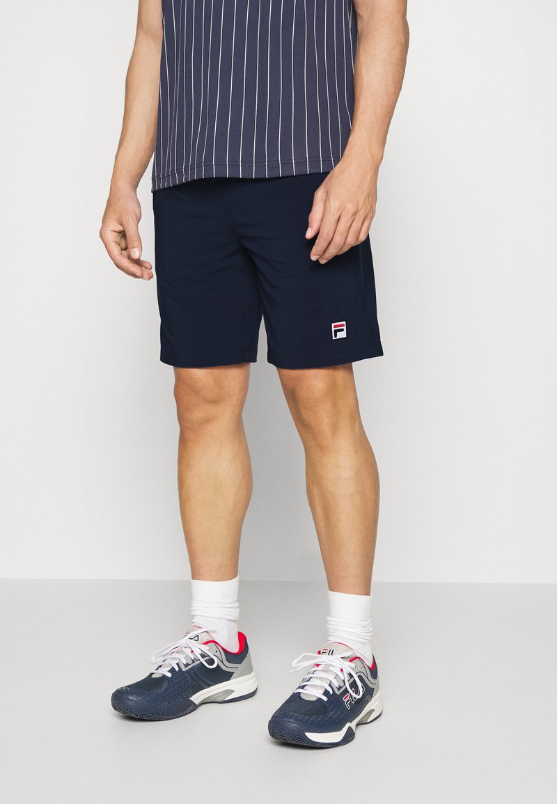Marine atletisk shorts med sidelommer og Fila-logo. Kombinert med en marineblå stripete kortermet topp og gråblå atletiske sko.