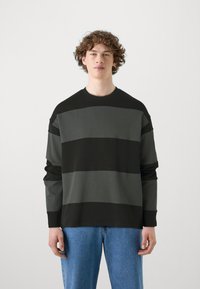 Mustmustriibuline musta ja halli värvi sweatshirt ümmarguse kaelusega ja pikkade varrukatega, kombineeritud siniste teksadega. Sile tekstuur, mugav lõige.