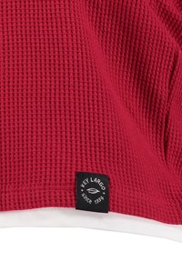 Tissu tricoté texturé rouge avec un ourlet blanc. Comprend une étiquette noire avec "KEY LARGO" et "depuis 1994" en blanc.