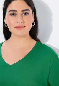 Zielona koszulka z dekoltem w serek, wykonana z miękkiego materiału, posiada krótki rękaw, gładką fakturę i luźne dopasowanie z delikatnym połyskiem. Widoczne srebrne kolczyki koła.