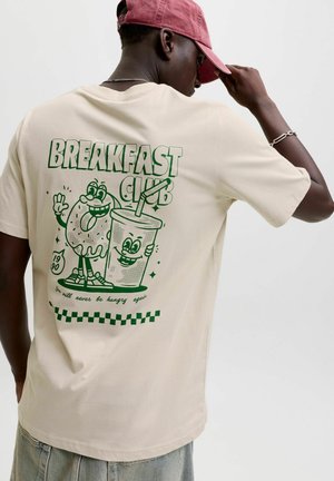 Mężczyzna ubrany w beżowy t-shirt z zielonym kreskówkowym pączkiem i napojem, tekst "BREAKFAST CLUB" i "Nigdy więcej nie będziesz głodny."
