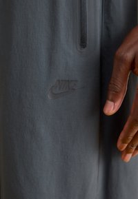 Pelēks Nike sporta tops ar puspīpējo dizainu. Iezīmēts ar teksturētu audumu un debossētu logo apakšējā kreisajā pusē. Redzamas rokas.