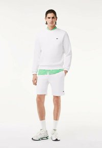 Felpa bianca e pantaloni corti con colletto verde e dettagli a toppa. Il modello indossa calzini bianchi e sneaker. Tessuto dalla texture liscia.