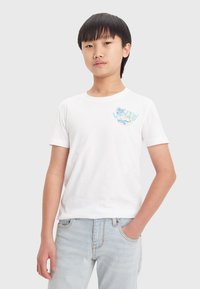Camiseta blanca de algodón con mangas cortas, que presenta un logo gráfico colorido en el lado izquierdo del pecho. Se lleva con unos jeans de mezclilla azul claros.