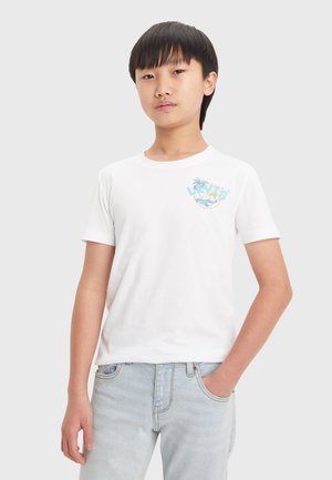 Camiseta blanca de algodón con mangas cortas, que presenta un logo gráfico colorido en el lado izquierdo del pecho. Se lleva con unos jeans de mezclilla azul claros.