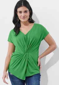 Grünes kurzärmliges Oberteil mit V-Ausschnitt und gedrehtem Vorderdesign, aus einem glatten, dehnbarem Material gefertigt. Kombiniert mit blauen Jeans.