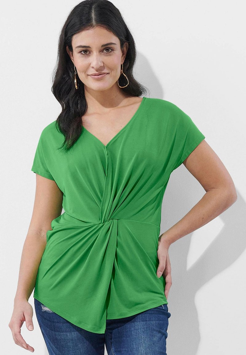 Grünes kurzärmliges Oberteil mit V-Ausschnitt und gedrehtem Vorderdesign, aus einem glatten, dehnbarem Material gefertigt. Kombiniert mit blauen Jeans.