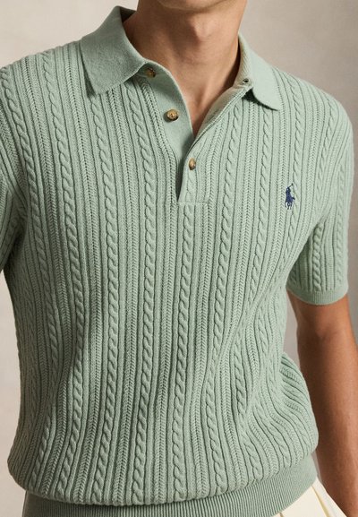 Homme portant un pull polo vert clair à manches courtes en maille torsadée avec trois boutons et un logo marine brodé sur la poitrine.