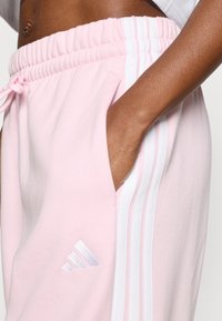 Rozā sporta bikses ar mīkstu tekstūru, baltām sānu svītrām un mazu sudrabotu logotipu kreisajā augšstilbā. Elastīga josta ar auklu.