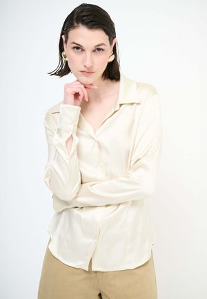 Giovane donna con capelli corti e scuri indossa una camicetta di seta color crema e pantaloni beige, poggiando il mento sulla mano e guardando la fotocamera con espressione neutra.