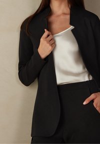 Czarna, dopasowana marynarka z wysokim kołnierzem, otwartym przodem i teksturowaną tkaniną, noszona na białym satynowym topie i czarnych spodniach z elastycznym pasem.