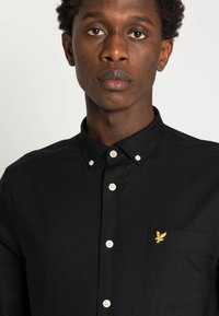 Μαύρο πουκάμισο με κουμπιά από βαμβάκι, διαθέτει τσέπη στην περιοχή του στήθους. Έχει λευκά κουμπιά και χρυσό κεντημένο λογότυπο στην τσέπη.