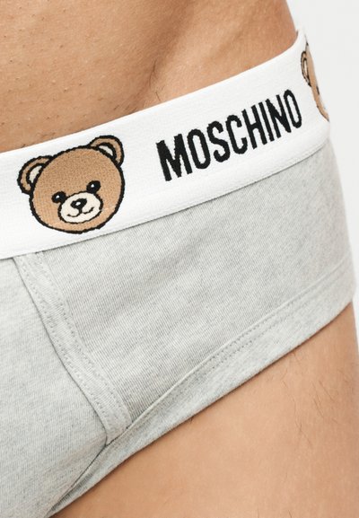 Κοντινό πλάνο γκρι εσώρουχου με λευκή μέση που φέρει πρόσωπα αρκουδάκια και το κείμενο «MOSCHINO», φορεμένο στη λεκάνη ενός ατόμου.