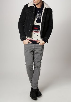 Giacca di jeans nera con colletto in pelliccia sintetica, maglione crema a fantasia, pantaloni grigi slim-fit e stivali neri alla caviglia. Outfit semplice e casual.