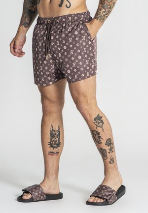 Mężczyzna w brązowych spodenkach w wzory i pasujących klapkach, prezentujący liczne tatuaże na nogach i ramionach, stojący na jednolitym tle.