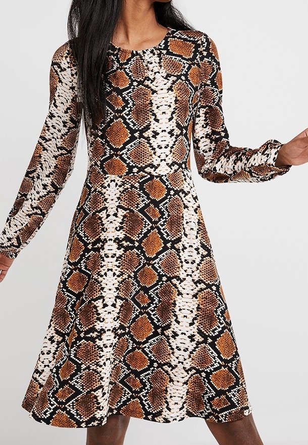 Femme portant une robe à manches longues, longueur genou, avec un motif serpent marron, noir et blanc sur un fond clair.