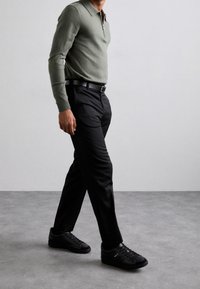 Polo verde a maniche lunghe con colletto zip, abbinato a pantaloni neri su misura e sneakers nere. Tessuto morbido, design aderente e stile minimalista.