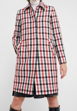 Femme portant un manteau mi-long à motif tartan rouge, noir et blanc, les mains dans les poches, par-dessus une jupe noire et un col roulé.