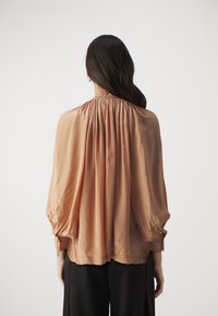 Peach-colored blouse made of lightweight fabric with a pleated back, loose-fit, and buttoned sleeves, paired with black trousers.