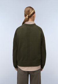Persona con capelli biondi raccolti, che indossa un maglione verde oliva sopra una camicia beige e pantaloni grigi, vista da dietro su uno sfondo neutro.