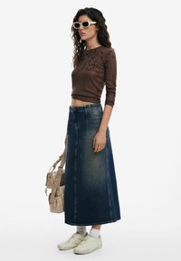 Džínová maxi sukně, modrá, vysoký pas, s předními švy; dlouhý hnědý pletený top s potiskem textu; krémové boty; béžová kabelka; bíl pozadí.