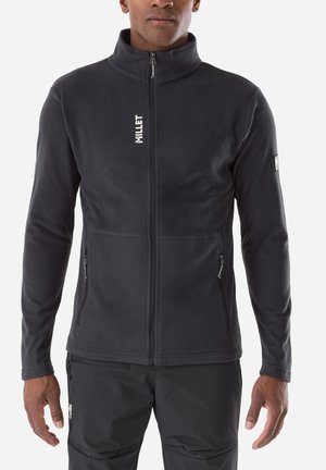 Homme portant une polaire zippée noire et un pantalon noir, debout avec les bras détendus le long du corps devant un fond uni.