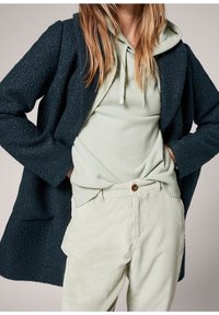 Tumma teksturoitu takki vaaleanvihreän hupparin päällä, yhdistettynä vaaleanvihreisiin samettihousuihin, joissa on nappikiinnitys ja näkyvät pystysuorat raidat.