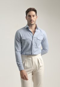 Camisa de manga larga azul claro con cuello abotonado, dos bolsillos en el pecho, combinada con pantalones plisados color crema. Textura de tela suave.