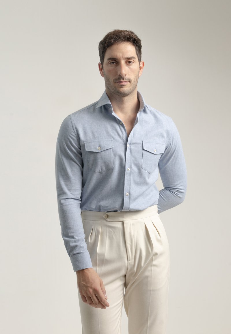 Camisa de manga larga azul claro con cuello abotonado, dos bolsillos en el pecho, combinada con pantalones plisados color crema. Textura de tela suave.