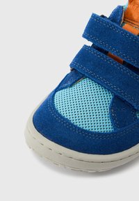 Μπλε σουέντ αθλητικό παπούτσι με ακροβατικά από δίχτυ σε ανοιχτό μπλε, με πορτοκαλί λεπτομέρειες και δύο λουράκια με βέλκρο. Λαστιχένια σόλα με υφή για καλύτερη πρόσφυση.