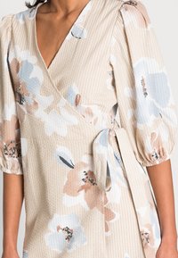 Krämfärgad omlottklänning med puffärmar, med ett blommönster i mjuk rosa, blå och svart. Strukturerad tyg med en knytning i midjan.