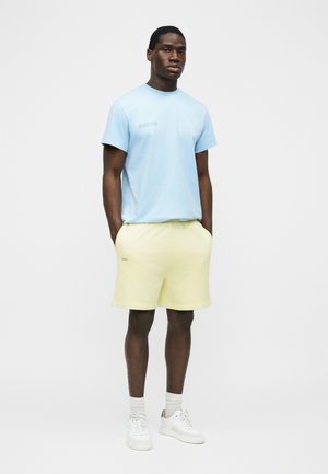 Uomo in piedi con le mani in tasca che indossa una maglietta azzurro chiaro, pantaloncini giallo pallido, calzini bianchi e scarpe da ginnastica bianche su sfondo neutro.