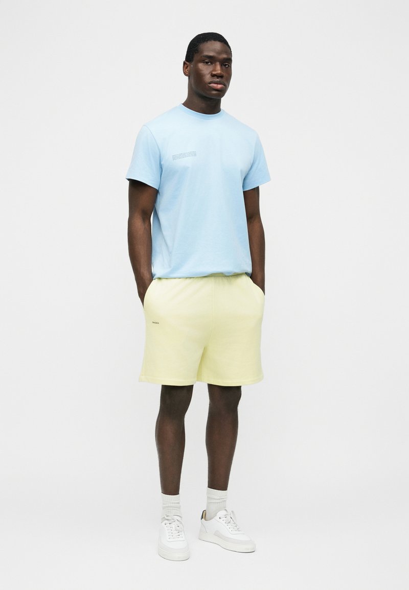 Man standing with hands in pockets wearing light blue t-shirt, pale yellow shorts, white socks, and white sneakers against a plain background.