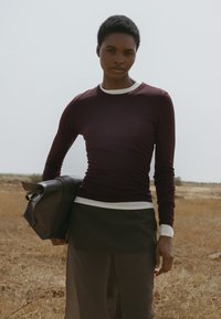 Maroonsidonnainen pitkähihaine paita valkoisella kauluksella, yhdistettynä tummiin hameeseen. Malli pitää tummasta nahkalaukusta, taustalla kuiva, avara maisema.