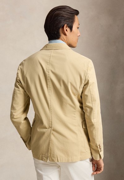 Homme vu de dos portant un blazer beige sur une chemise bleu clair et un pantalon blanc sur un fond neutre uni.