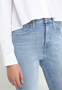 Primer plano de una persona que lleva unos pantalones vaqueros de tiro alto de color azul claro y una camisa blanca de manga larga, con la mano descansando a un lado.