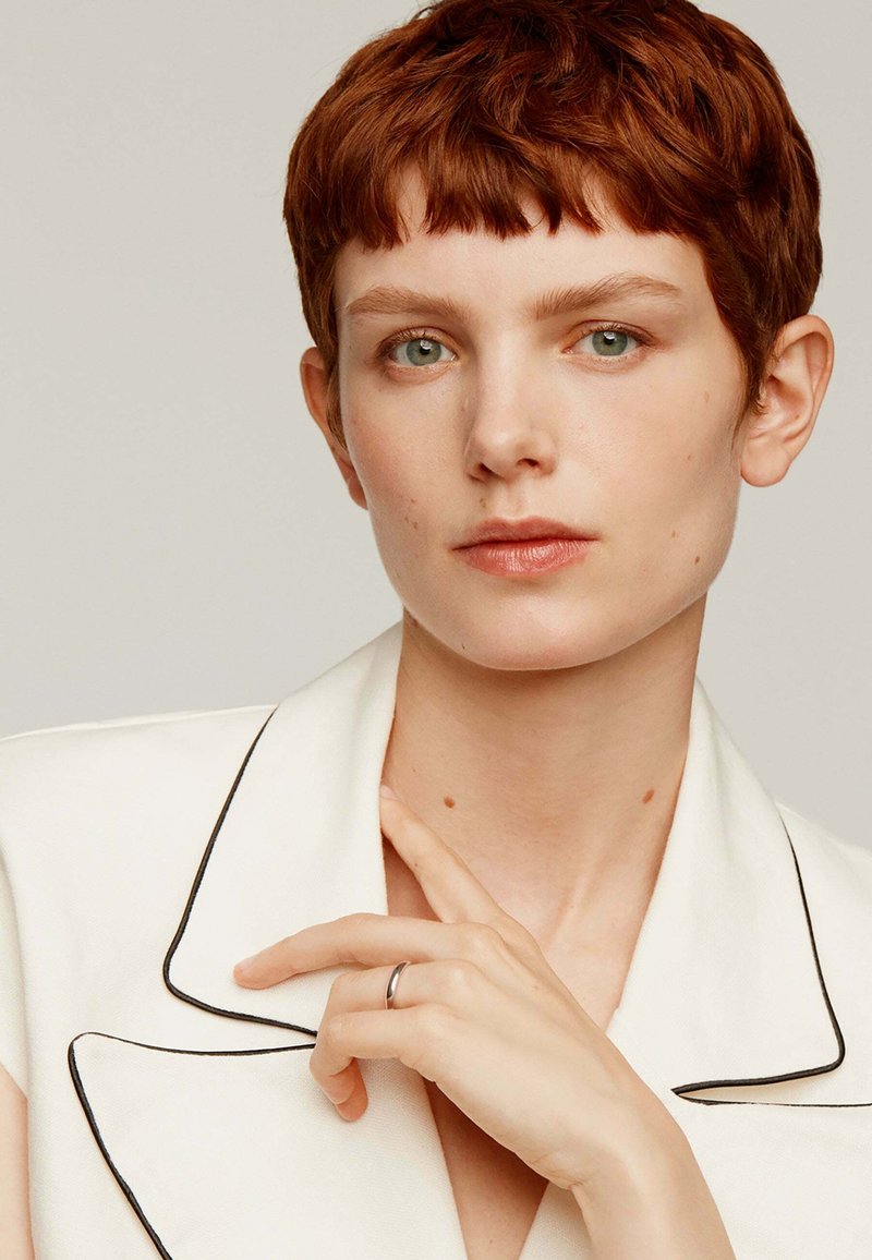 Blazer blanco con ribetes negros, cabello corto rojo, maquillaje neutro y anillo de plata en el dedo. Fondo limpio y mirada directa.