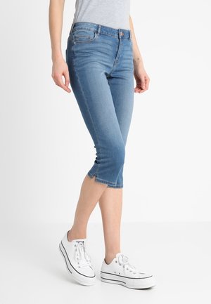 Personne portant un jean en denim bleu clair court et des baskets blanches à lacets, debout devant un fond uni clair.