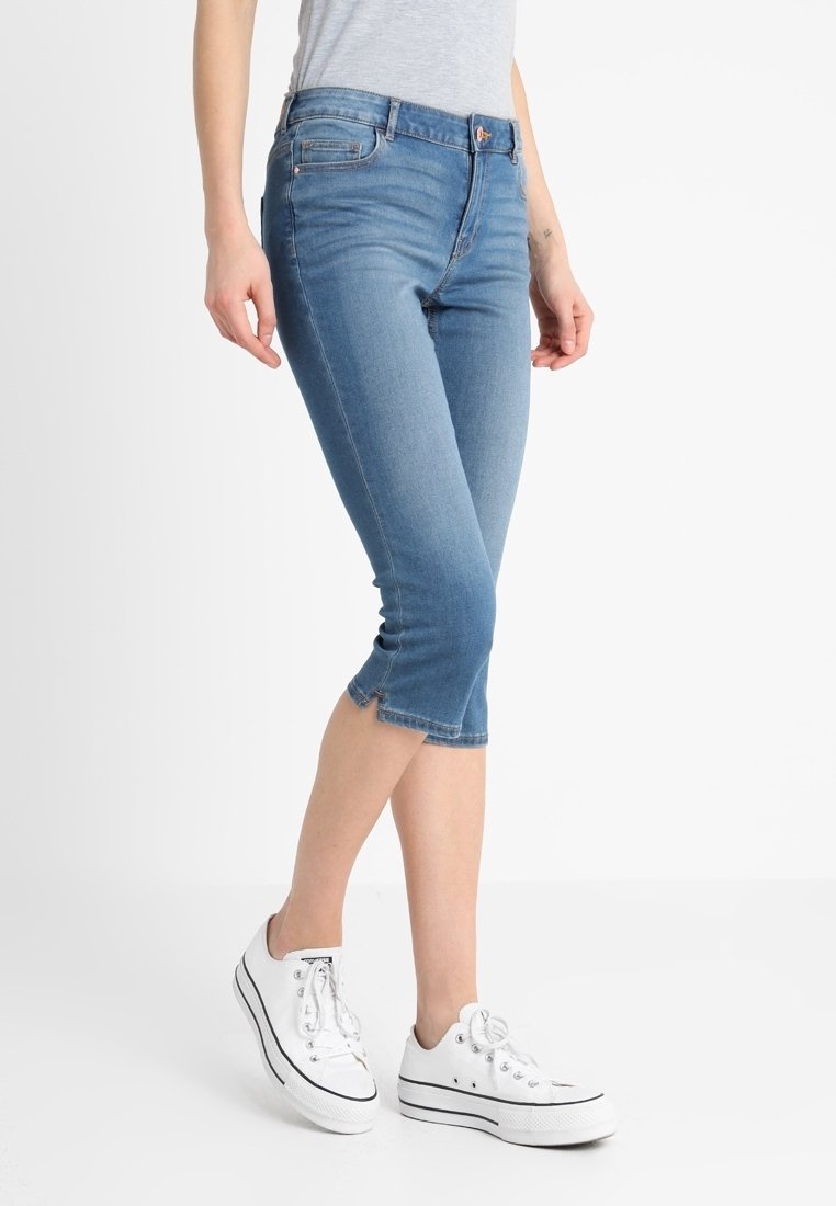 Personne portant un jean en denim bleu clair court et des baskets blanches à lacets, debout devant un fond uni clair.