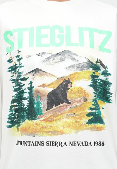 Biela bavlnená tričko s grafikou medveďa na klade medzi zelenými stromami a horami. Text znie "STIEGLITZ" a "MOUNTAINS SIERRA NEVADA 1988."