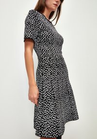Schwarzes Kleid mit weißen Punktmustern, kurzen Ärmeln, tailliertem Oberteil, ausgestelltem Rock, weichem Stoff und runder Halslinie.