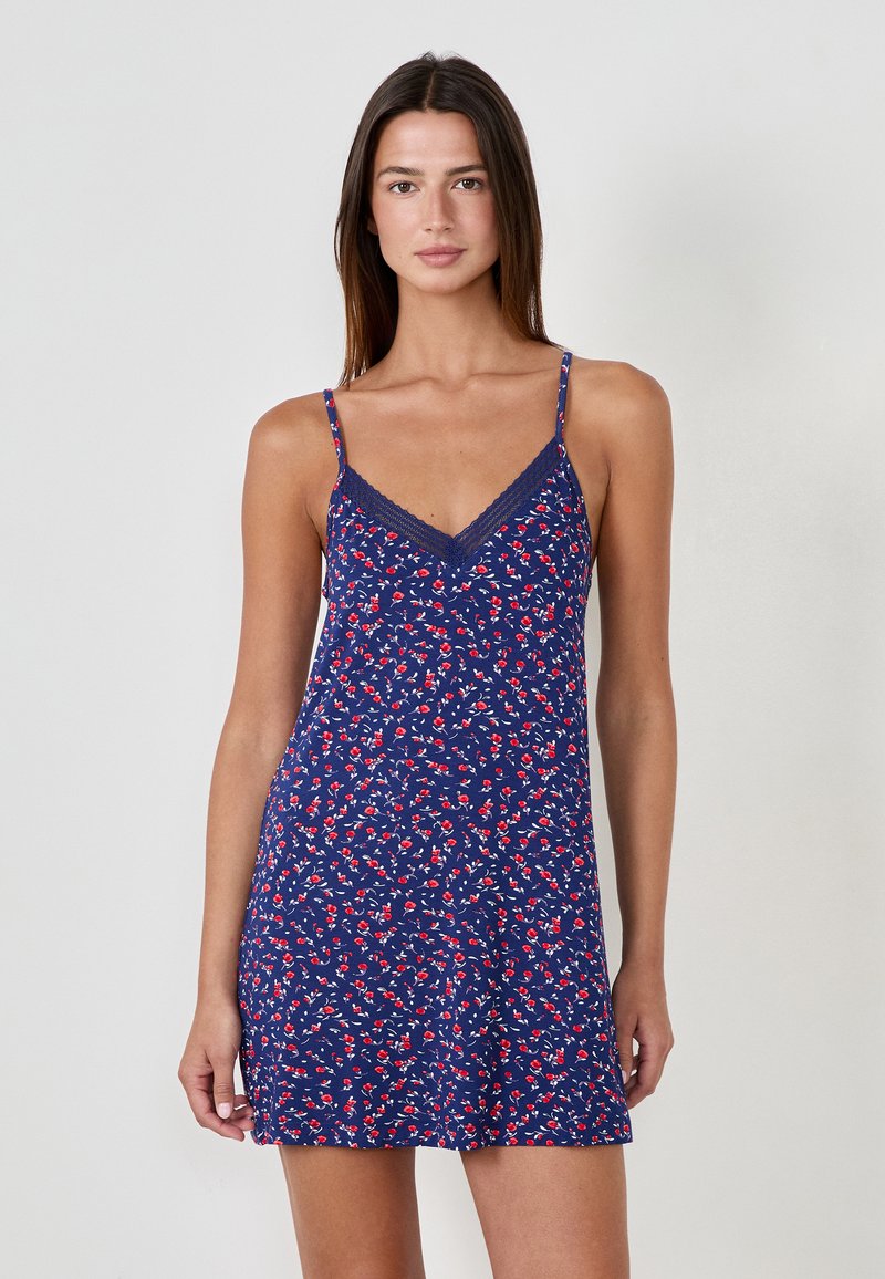 Robe nuisette marine avec imprimé floral rouge et blanc. Elle dispose de bretelles fines et d'une bordure en dentelle au niveau du décolleté. Tissu léger et coupe décontractée.