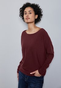 Bordeaux langemouwen trui met een wijde halslijn en gerimpelde manchetten, gecombineerd met donkerblauwe jeans, op een lichtgrijze achtergrond.