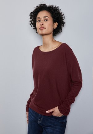 Un pull bordeaux à manches longues avec un col large et des poignets froncés, associé à un jean bleu foncé, sur un fond gris clair.