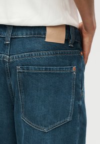 Μπλε τζιν παντελόνι σε άτομο, εμφανίζοντας την πίσω τσέπη, τις θηλιές της ζώνης και μια ταμπέλα από δέρμα καφέ χρώματος στη μέση.