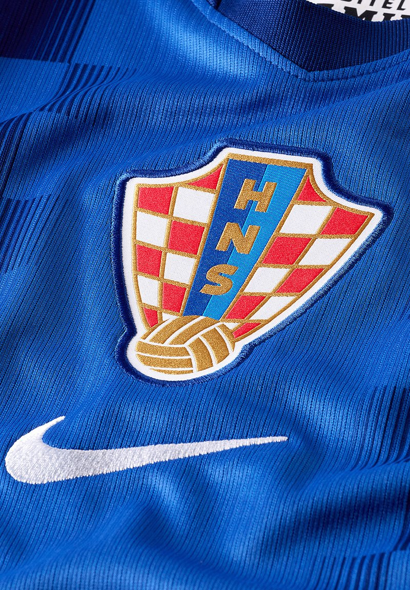 Camiseta deportiva azul con un escudo a cuadros rojo y blanco con las letras "HNS" y un balón de fútbol dorado, junto a un logo blanco de Nike.