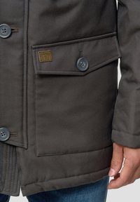 Manteau marron avec grande poche rabat boutonnée et coutures visibles, porté par une personne en jean bleu, main détendue sur le côté.