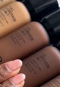 Tre flasker med "FULL COVERAGE 2-IN-1" foundation og concealer i forskellige hudtoner, med sorte kapsler og præget etiketter.